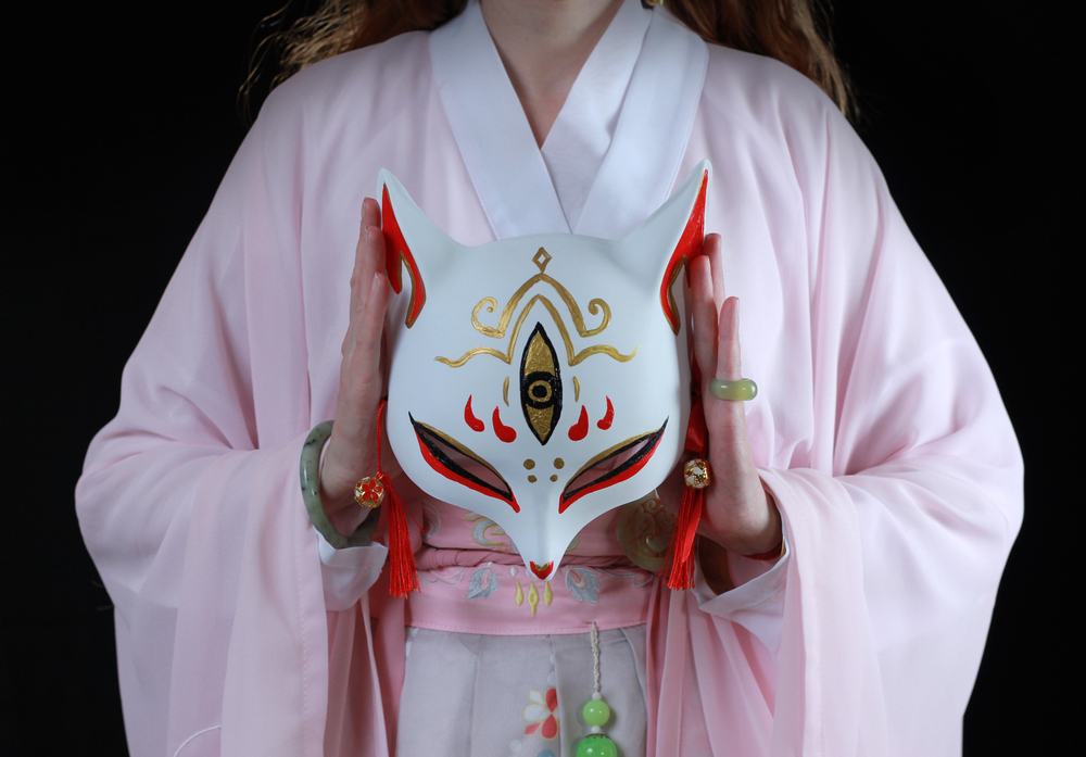 ผู้หญิงสวมชุดกิโมโนหรือฮันฟูและหน้ากากจิ้งจอก หน้ากากจิ้งจอกญี่ปุ่นแบบดั้งเดิมที่เรียกว่าคิตสึเนะ แนวคิดดั้งเดิมของจีนและญี่ปุ่น ตำนานมหัศจรรย์