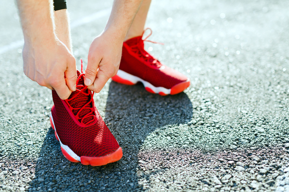 คอนเซ็ปต์สปอร์ตรองเท้าผ้าใบสีแดงสำหรับการวิ่ง ผู้ชายกำลังผูกเชือกรองเท้าผ้าใบโดยไม่มีใบหน้า ขาในรองเท้ากีฬามืออาชีพปิดสนิท กลางแจ้ง แสงแดด สนามกีฬา